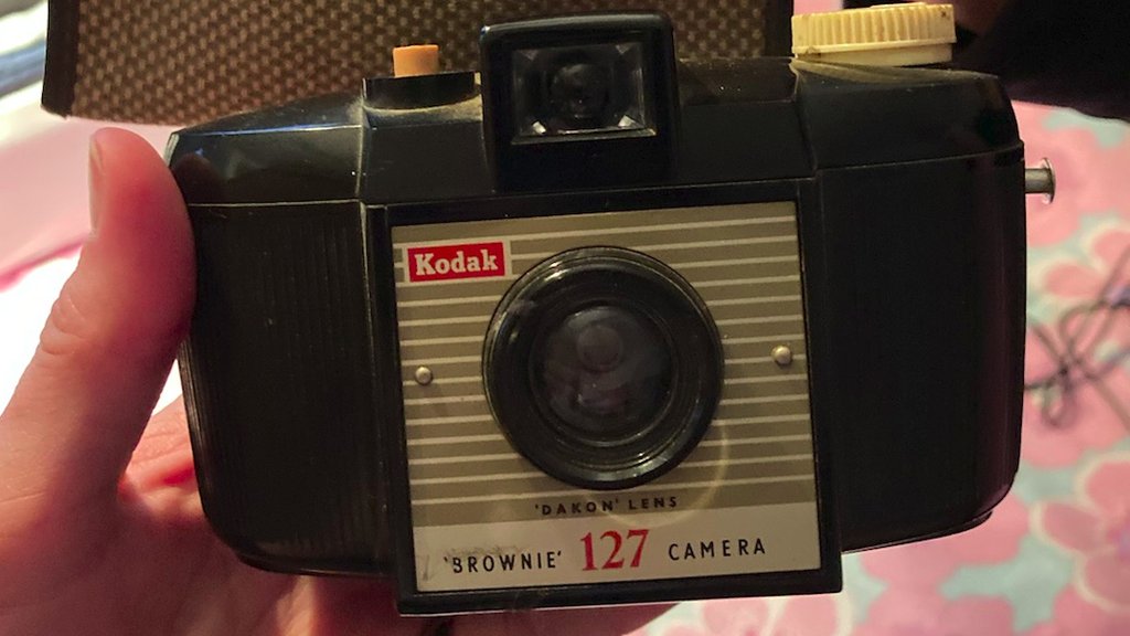 Das Bild zeigt eine Kodak Brownie 127 Kamera. Die Kamera ist schwarz-braun mit einem grau-beige gestreiften Bereich, auf dem der Name "Kodak" und die Bezeichnung "Brownie 127 Camera" zu lesen sind. Das Objektiv ist sichtbar, und ein Teil des Kameragehäuses scheint abgenutzt zu sein.