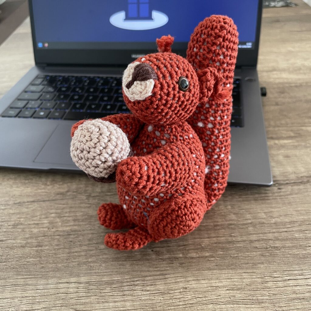 Das Bild zeigt einen handgefertigten, rot-braunen Häkel-Eichhörnchen, das eine beigefarbene Nuss in seinen Vorderpfoten hält. Das Eichhörnchen sitzt auf einem Holztisch vor einem Laptop, dessen Bildschirm im Hintergrund unscharf ist.  Die Szene wirkt gemütlich und heimelig.
